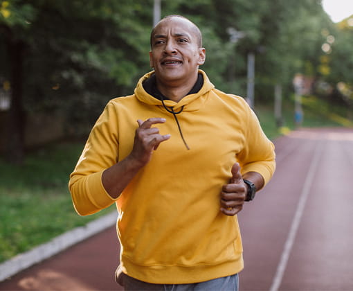 Un hombre viste una sudadera mientras corre entusiasmado en una pista de atletismo rodeada por arboles frondosos.