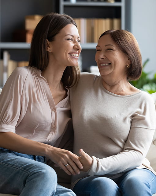 Dos mujeres sentadas en la sala de un hogar. Ellas sonríen con mucha alegría bajo la agradable luz que entra por la ventana.
