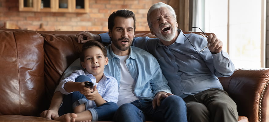 Un niño está sentado junto a su padre y su abuelo en un cómodo sofá. Los tres se están riendo de lo que ven en el televisor.
