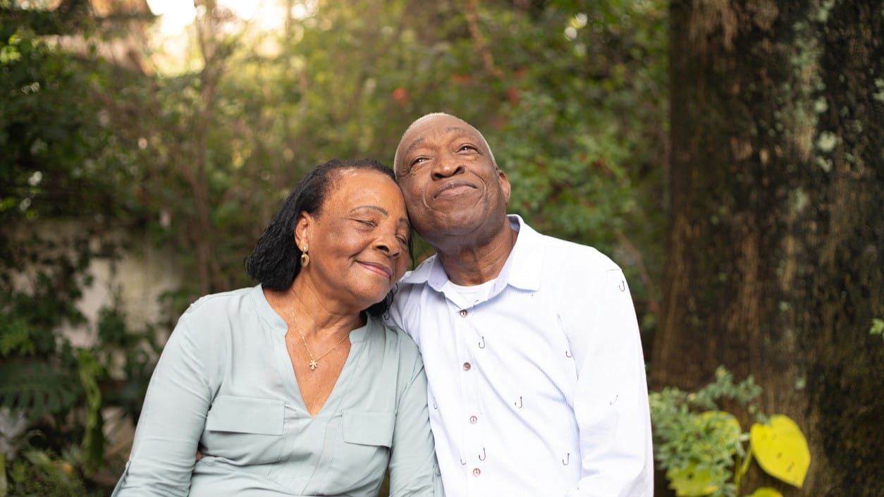  An older couple in a wooded setting, gently leaning into each other.