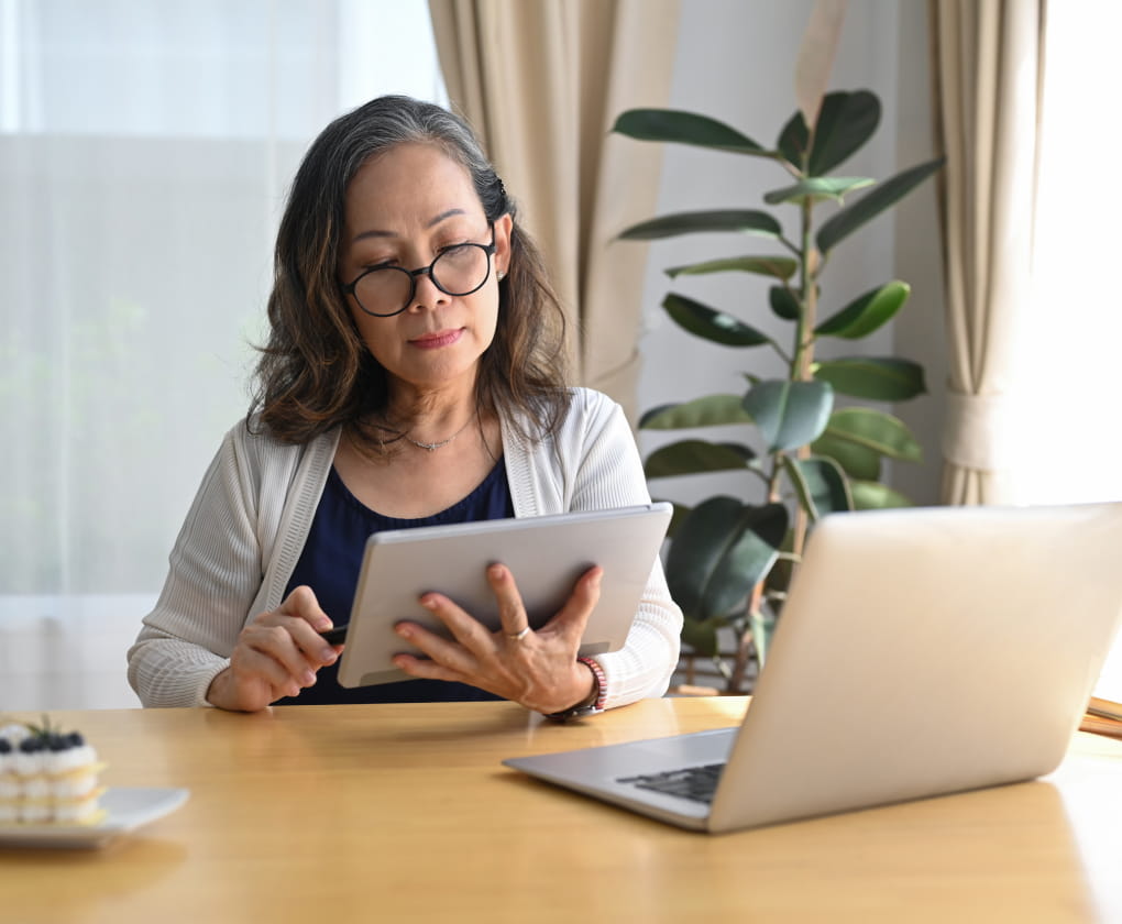 A woman reads from her tablet with a laptop in front of her.