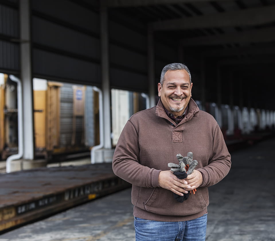 A smiling man standing by train tracks holding a pair of gloves.