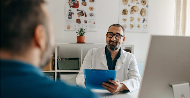 A healthcare provider seated at a desk holding a clipboard and talking to a man