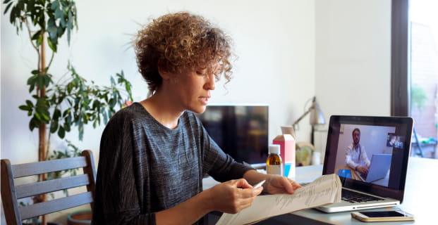 A woman holding paperwork and sitting in front of a laptop, talking to a doctor who is visible on the laptop screen