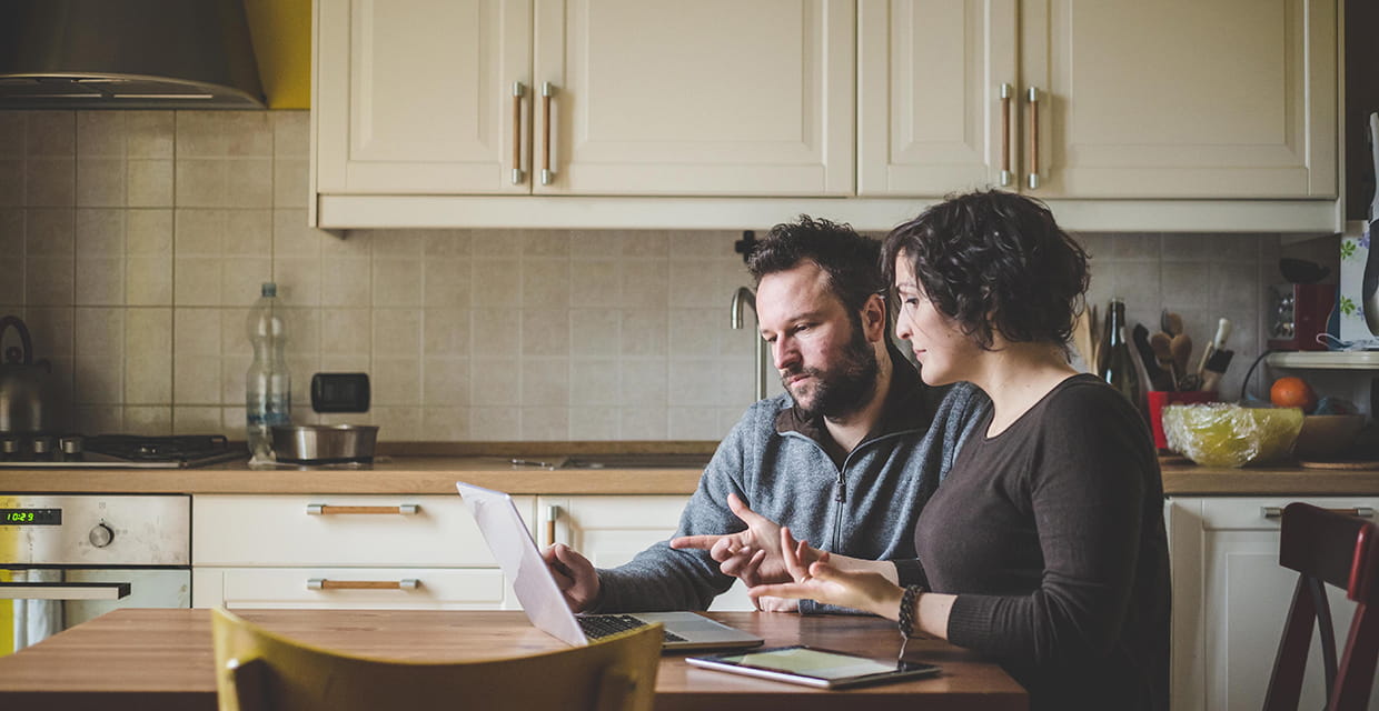 A man and woman sitting at the kitchen table looking at a laptop.