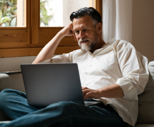 A man with a beard sits in a chair, leaning his head on one fist, looking at a laptop computer in his lap.