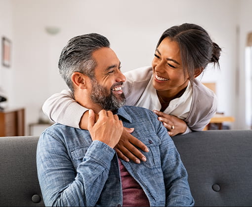 Una mujer y un hombre sonríen mientras se miran a los ojos. Ella lo abraza mientras él está sentado en un sofá agarrando una de las manos de ella.