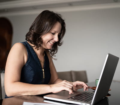 Una mujer está sentada en la mesa de comer de su casa. Ella escribe un mensaje en su laptop acompañada por una taza de café.