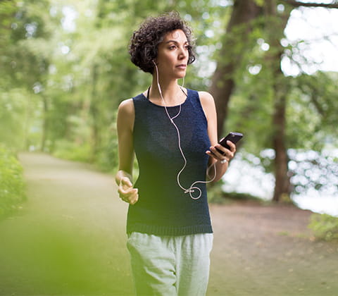 Una mujer pasea con audífonos puestos en un camino junto a un lago y rodeada por naturaleza muy verde.